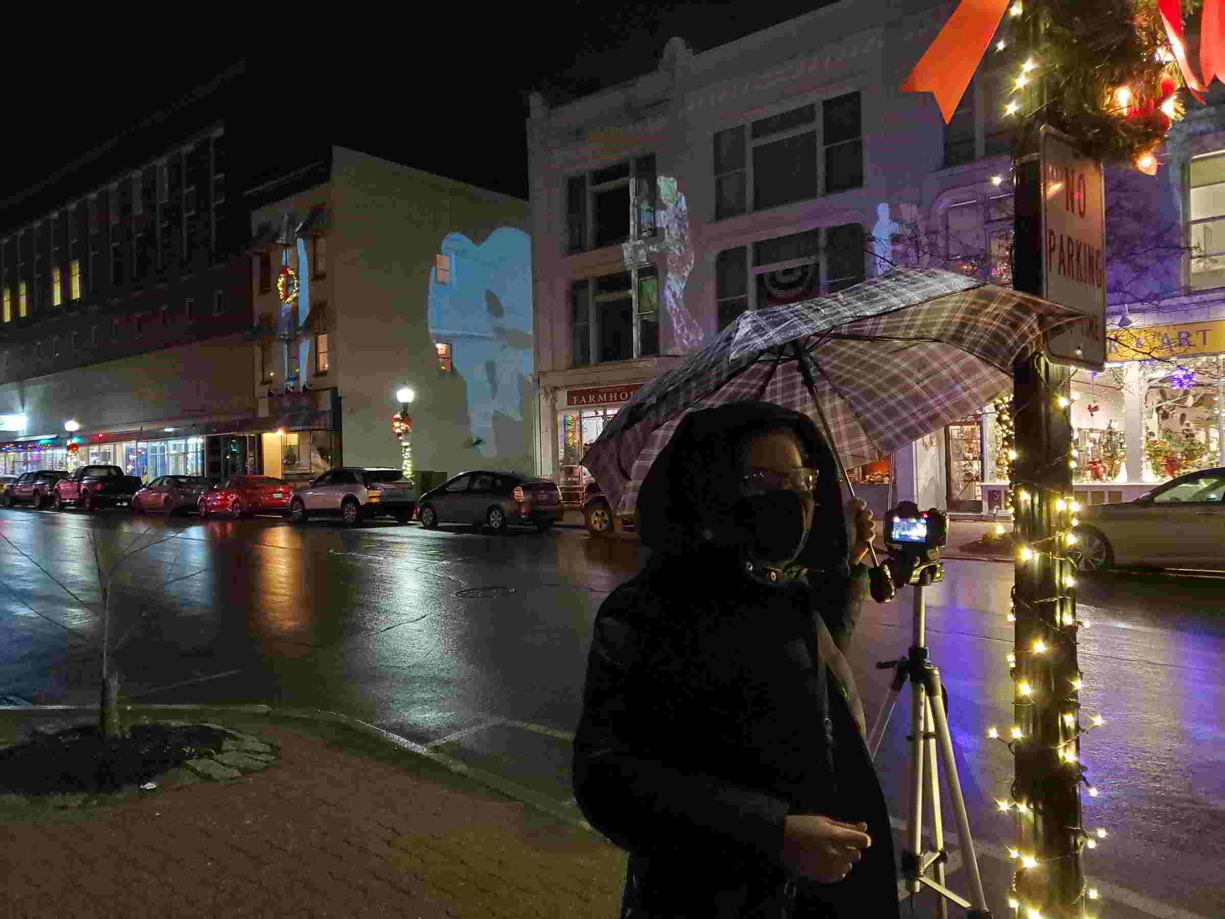 Katarina in winter jacket with umbrella in front of camera stand with pictures of dancers projected across the street.