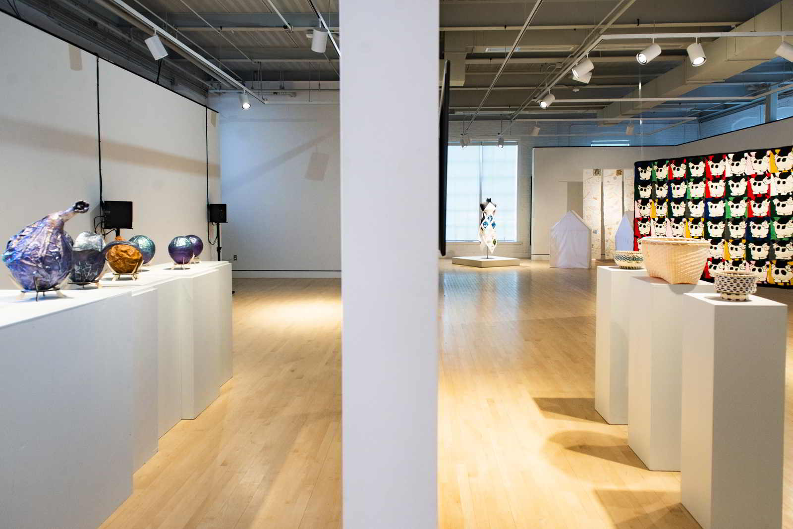A photo of the empty Lord Hall Gallery with all 2022 MFA thesis projects visible. On the left hand side, purple spherical shapes and an empty space surrounded by black speakers can be seen. On the right, baskets, a dress, houses, and a hanging rug can be seen.  