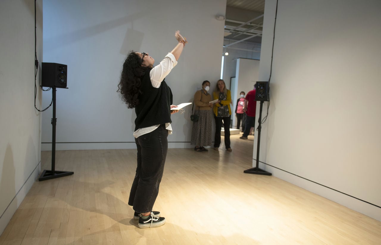 A lady stands in the middle of the gallery space, stretching up with her hands towards the ceiling.